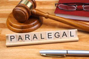 Wooden Scrabble tiles spelling out 'PARALEGAL' with gavel, book, glasses in background and a pen in foreground.